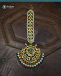 A close-up image of a Hyderabadi Jadau maang tika, adorned with green and white CZ Polki stones and faux pearls. The maang tika is handcrafted with gold-plated alloy and features intricate Jadau work.