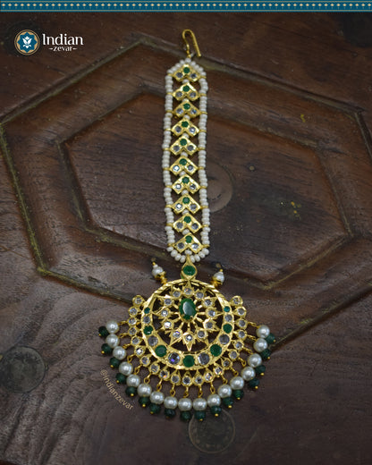 A close-up image of a Hyderabadi Jadau maang tika, adorned with green and white CZ Polki stones and faux pearls. The maang tika is handcrafted with gold-plated alloy and features intricate Jadau work.