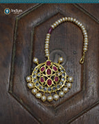 A close-up image of a Hyderabadi Jadau maang tika, adorned with rubies, CZ Polki stones, and real freshwater pearls. The maang tika is handcrafted with gold-plated alloy and features intricate Jadau work