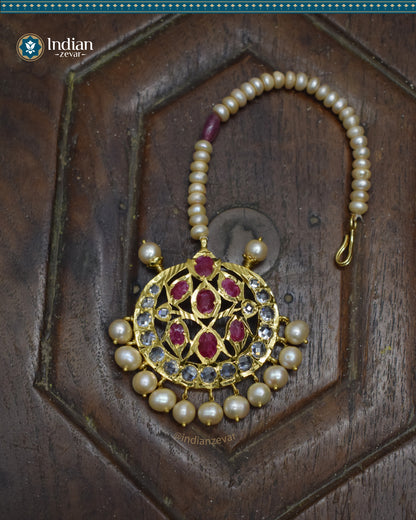 A close-up image of a Hyderabadi Jadau maang tika, adorned with rubies, CZ Polki stones, and real freshwater pearls. The maang tika is handcrafted with gold-plated alloy and features intricate Jadau work