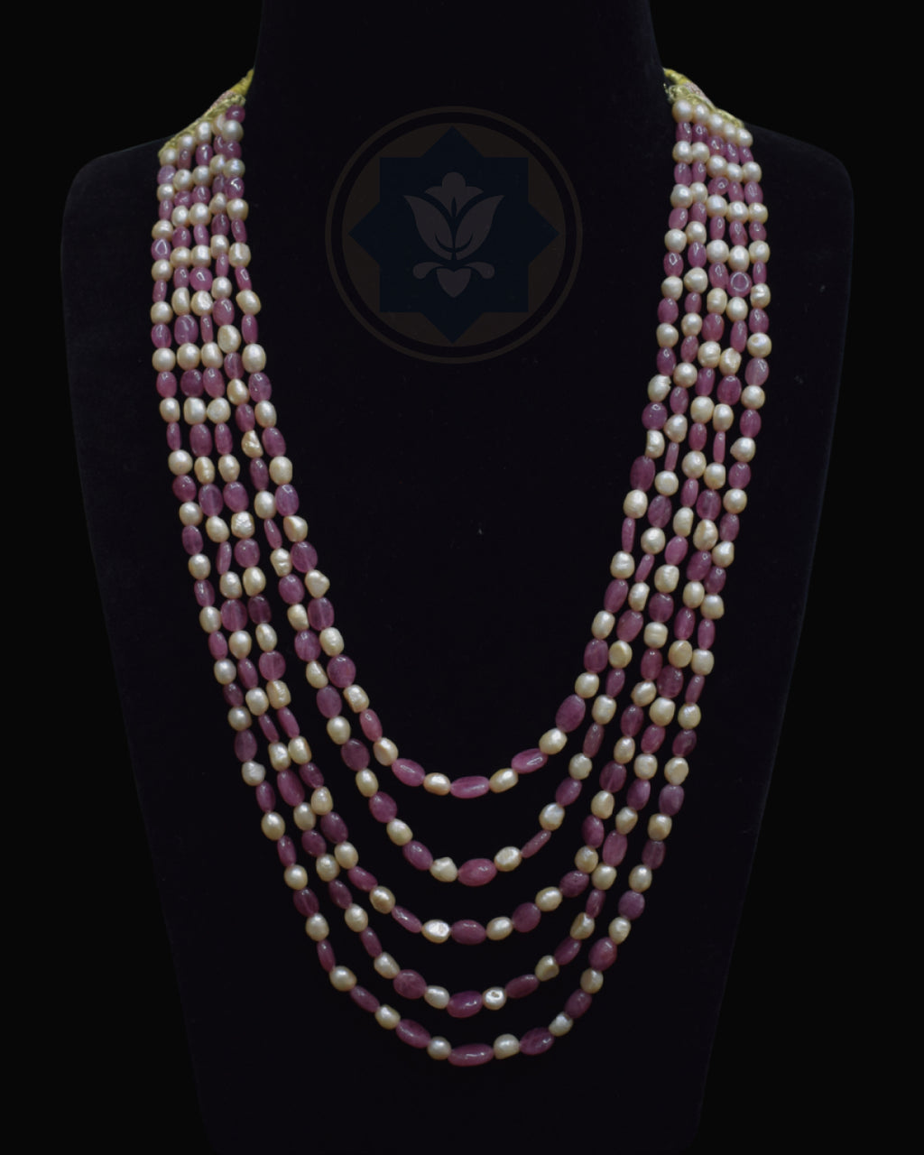 A multi-strand necklace displayed on a black mannequin bust against a black background. The necklace features alternating strands of faceted dusty rose ruby beads and cream-colored freshwater pearls, with a dark blue geometric logo printed on the center of the bust.
