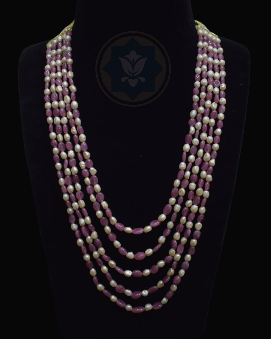 A multi-strand necklace displayed on a black mannequin bust against a black background. The necklace features alternating strands of faceted dusty rose ruby beads and cream-colored freshwater pearls, with a dark blue geometric logo printed on the center of the bust.