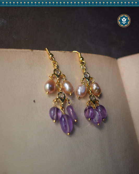 The image shows a pair of gold-plated metal earrings with purple amethyst beads and white pearls. The earrings are in a hoop style with a dangle drop. The amethyst beads are round and the pearls are oval. The earrings are sitting on top of a book.